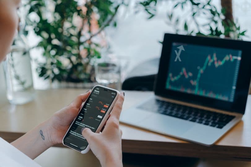 Mains tenant un smartphone affichant des cours, devant un ordinateur portable montrant un graphique en chandeliers